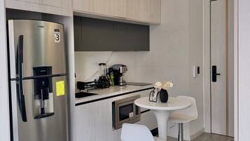 Una cocina moderna con un refrigerador de acero inoxidable, un microondas integrado, una cafetera y un dispensador de agua. Hay una mesa redonda pequeña con dos taburetes blancos.