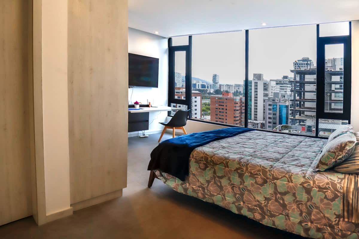 Un dormitorio moderno con una cama grande, un escritorio y una silla, que ofrece vistas a la ciudad a través de amplias ventanas.