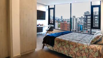 A modern bedroom with a large bed, a desk, and a chair, featuring a city view through large windows.