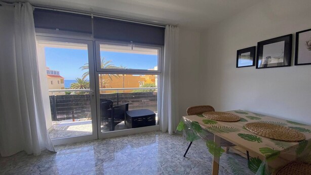 A room with a sliding glass door leading to a balcony, a table with a leaf-patterned tablecloth, and two wicker chairs. - Apt. ManRey 2A - Turquesa (Valle Gran Rey)