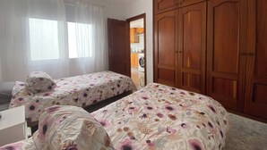 A bedroom with two beds, a wooden wardrobe, a window with sheer curtains, and a view into a kitchen area. - Apt. ManRey 2A - Turquesa (Valle Gran Rey)