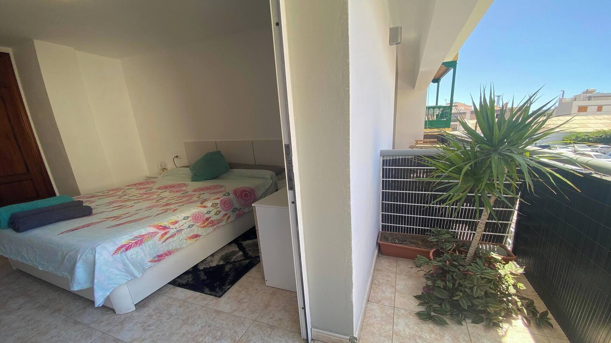A bedroom with a bed, a nightstand, and a window. There is a balcony with a railing and a potted plant.