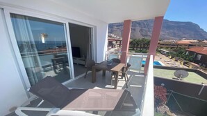Un balcon avec une table et des chaises, un téléviseur, et une vue sur la montagne et la piscine.