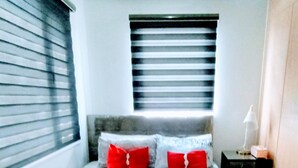 Una cama bien hecha con almohadillas rojas, una lámpara de noche y una ventana con persianas.