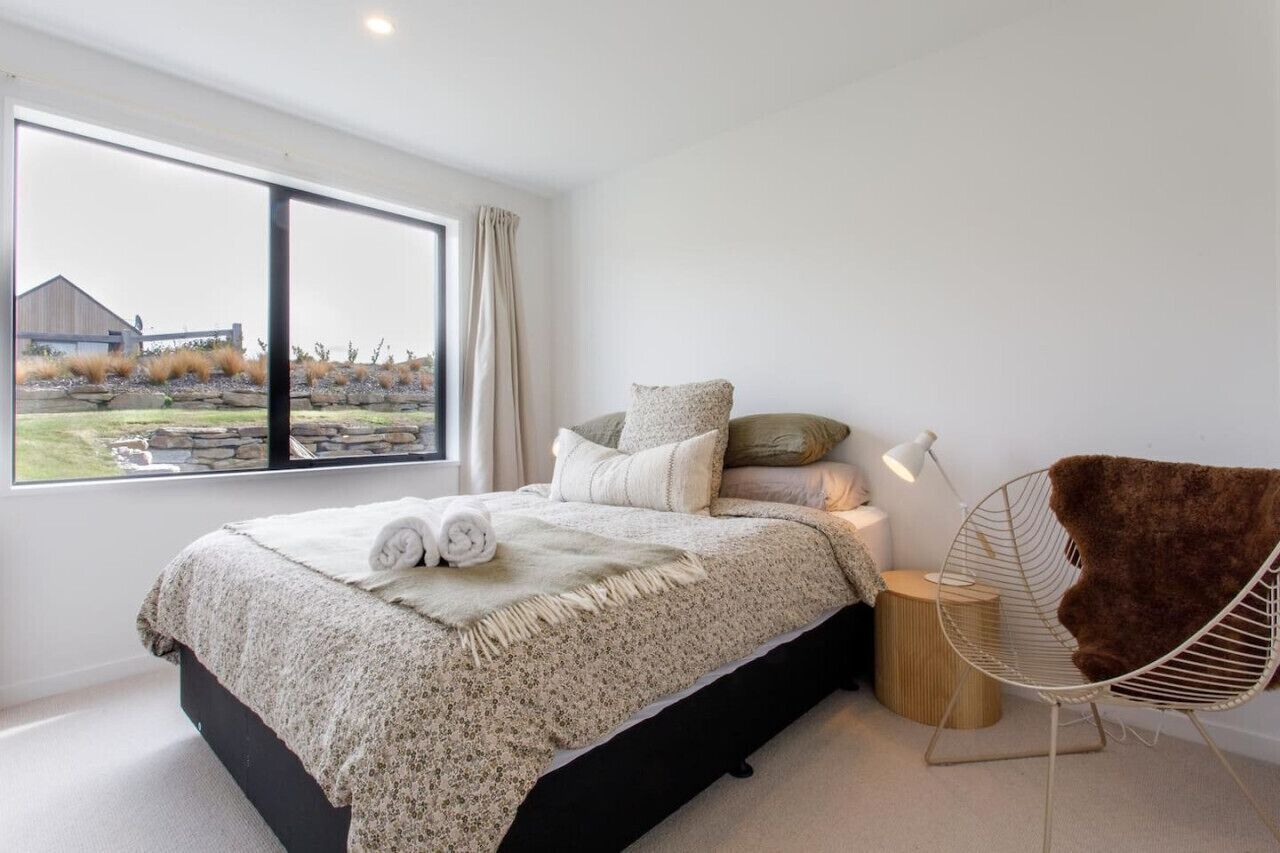 A modern bedroom with a large window, a bed with a white and gray patterned bedspread, a fluffy brown chair, and a small bedside table with a lamp.