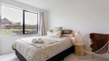 A modern bedroom with a large window, a bed with a white and gray patterned bedspread, a fluffy brown chair, and a small bedside table with a lamp.