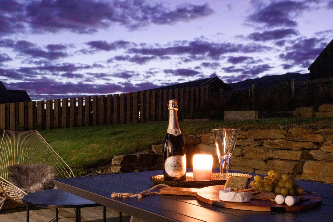 A table set with a bottle of wine, a glass, and a cheese board with grapes and crackers, outdoors at dusk.