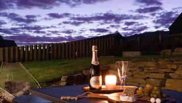 A table set with a bottle of wine, a glass, and a cheese board with grapes and crackers, outdoors at dusk.