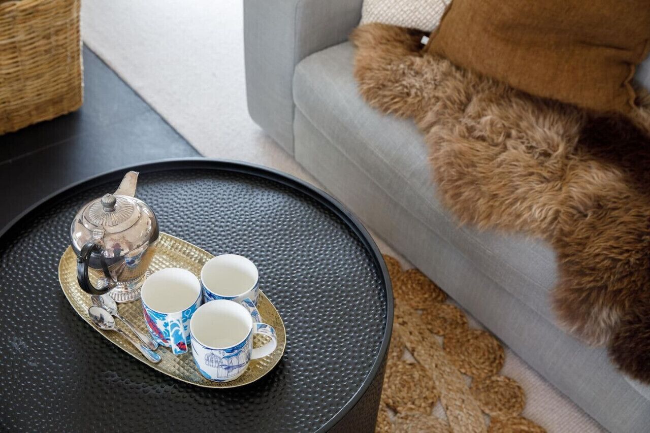 A tea set on a tray with a teapot and cups on a round table next to a sofa with a furry throw.