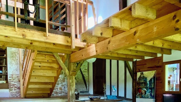 Een ruime woonkamer met een gele bank, houten balken, een trap en uitzicht op het landschap.