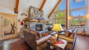 A cozy living room with a stone fireplace, leather sofas, and large windows offering a view of the outdoors.