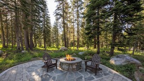 A circular stone fire pit with two wooden Adirondack chairs facing it, surrounded by a paved patio.