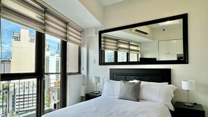 Habitación de hotel con una cama grande, dos mesitas de noche, un tocador, un espejo y vistas de la ciudad a través de ventanas de pared a techo.