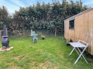 Een gazon met een houten tuinhuis, een metalen tafel, twee metalen stoelen, een zwarte pot met bloemen en een metalen kooktoestel.