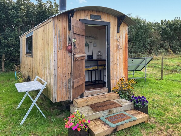 A wooden cabin with a door open, revealing a kitchen area with a sink and cabinets, and a small outdoor seating area with a table and chairs.