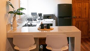 Een kleine keuken met een wit aanrecht, twee witte barkrukken, een zwarte koelkast en een houten plafond.