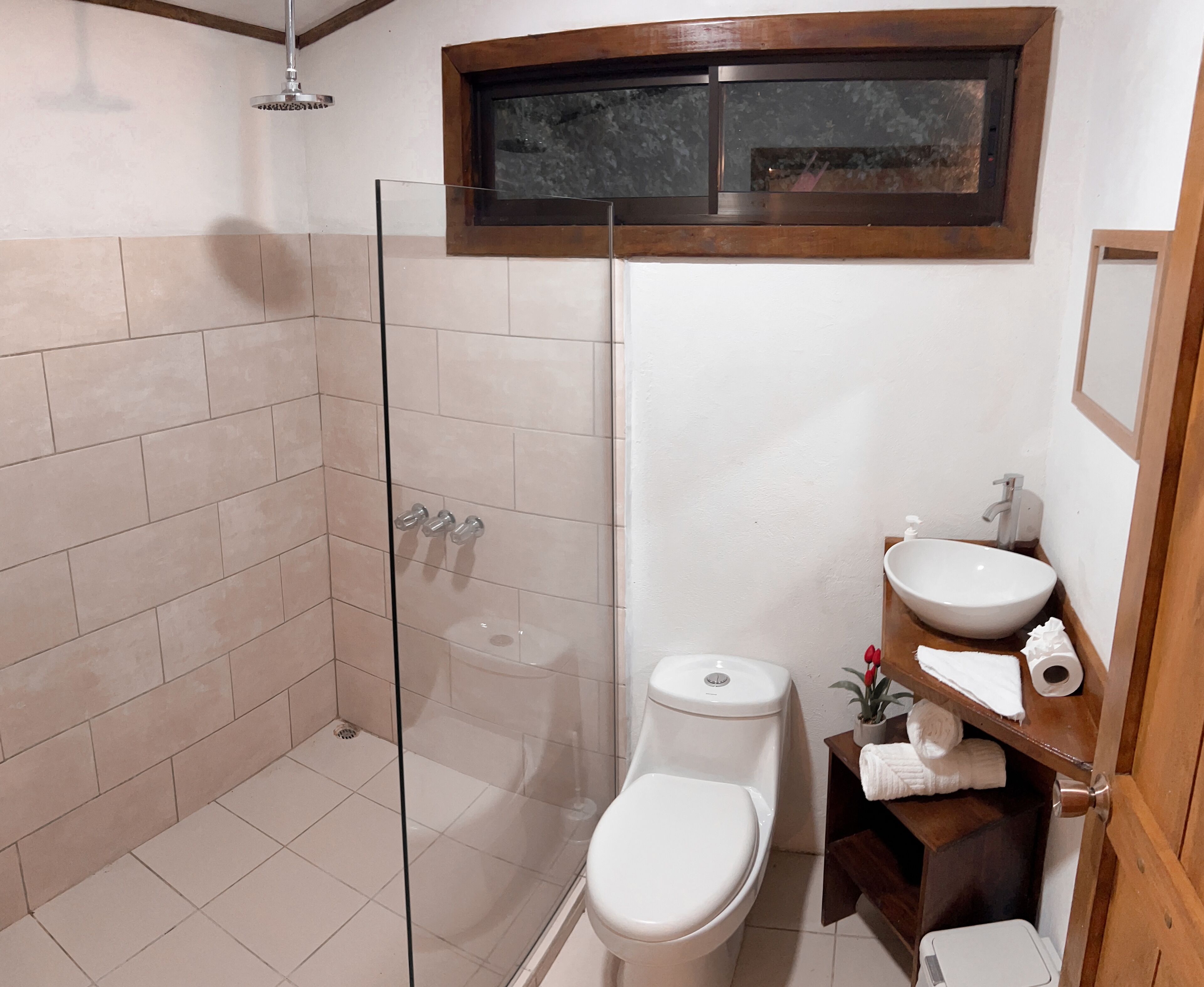 Un baño con ducha de vidrio, inodoro, lavamanos y un pequeño estante con toallas.