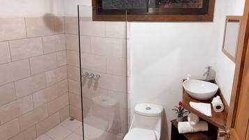 Un baño con ducha de vidrio, inodoro, lavamanos y un pequeño estante con toallas.