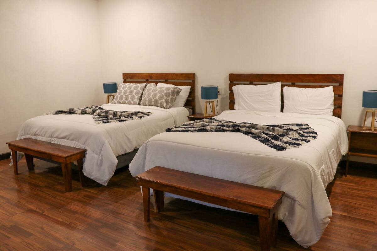 Ein Hotelzimmer mit zwei Einzelbetten, jeweils mit Holz-Kopfteil, weißer Bettwäsche und einem gemusterten Überwurf. Zwei Nachttische mit Lampen und eine Holzbank vor den Betten.