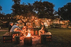 Un groupe de personnes est assis autour d’un feu de camp dans un jardin, avec une maison en arrière-plan.