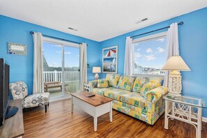 A room with a blue wall, a sofa with colorful cushions, a wooden coffee table, a TV, and a balcony with a view.