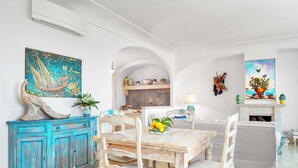 Comedor con mesa de madera, sillas blancas y un mueble azul. Hay un cuadro en la pared y un estante con adornos.