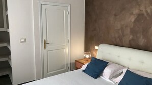 A bedroom with a white bed, two blue pillows, a white headboard, a nightstand with a lamp, and a door with a handle.