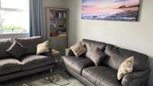 A living room with two grey sofas, a bookshelf, and a painting of a sunset over the sea. - Birdsong View - comfortable apartment in Amble (Amble)
