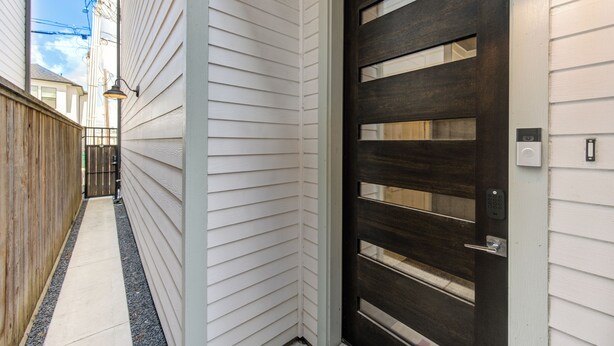 Un estrecho sendero conduce a una puerta de madera oscura con cerradura electrónica, revestimiento blanco en la casa y un pequeño aplique exterior.