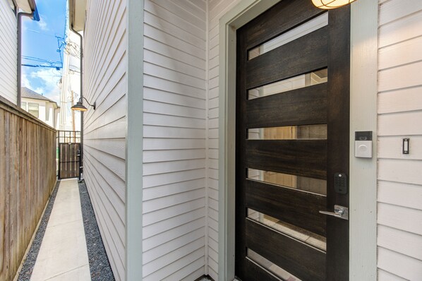 Un estrecho sendero conduce a una puerta de madera oscura con cerradura electrónica, revestimiento blanco en la casa y un pequeño aplique exterior.