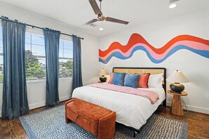 Habitación de hotel con cama, cortinas, ventilador de techo, un mural en la pared, lámparas de noche y un banco.