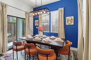 Comedor con una mesa dispuesta para seis personas, sillas naranjas, un florero con flores y una pared azul con dos ventanas y cortinas.