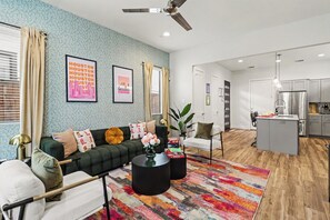 Una sala moderna con un sofá, una mesa de centro y un tapiz colorido. Cuenta con una zona de cocina con refrigerador y una mesa de comedor.