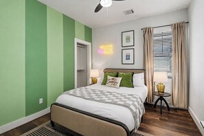 A bedroom with a bed, bedside lamps, a neon sign, a window with curtains, and a door.