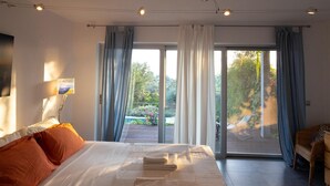 A modern bedroom with a large bed, a view of a garden through a sliding glass door, and a seating area with a sofa. - Villa Milos - Three Bedroom Villa, Sleeps 6 (Lefkada)