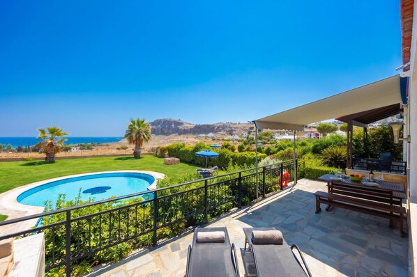 A pool with a view of the sea, a patio with a table and chairs, and a covered area with a grill.