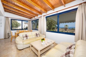 A room with a wooden ceiling, a sofa, a dining table, and large windows with a view of the outdoors.