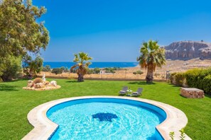 A swimming pool with a view of the sea, surrounded by grass and palm trees.