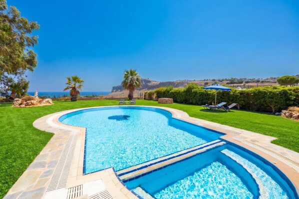 A swimming pool with a curved edge, surrounded by a well-maintained lawn and a view of the sea.