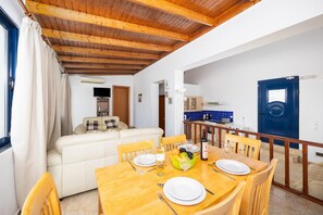 A dining area with a wooden table set for four, a sofa in the background, and a kitchen visible through an open door.
