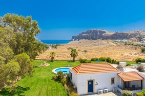 A residential property with a swimming pool, a well-maintained lawn, and a view of the sea and a rocky hill in the background.