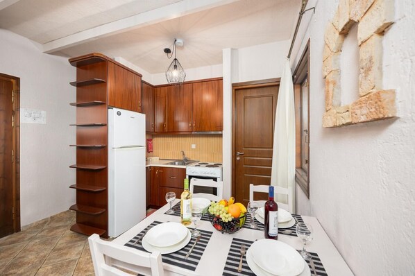 A compact kitchen with wooden cabinets, a white refrigerator, a stove, and a dining table set for four with plates, glasses, and a fruit bowl. - Panorama II Villas - Two Bedroom Villa, Sleeps 7 (Ditiki Mani)