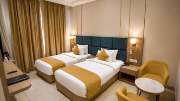 A hotel room with two beds, a desk, a chair, a small table, and a ceiling fan.