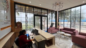 A hotel reception area with a receptionist at a desk, large windows offering a view of the outdoors, and a seating area with sofas. - Hotel Three Seasons (Panigaon)