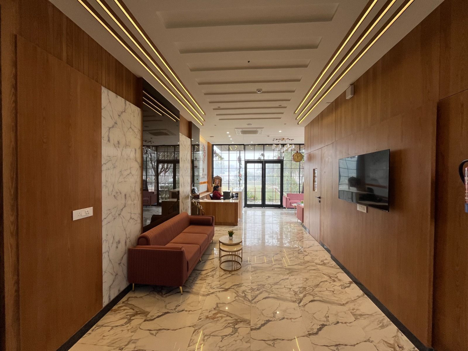Um lobby de hotel moderno com piso de mármore, painéis de madeira, um sofá, uma mesinha redonda e uma TV fixada na parede.