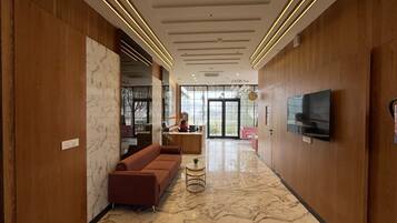 Um lobby de hotel moderno com piso de mármore, painéis de madeira, um sofá, uma mesinha redonda e uma TV fixada na parede.