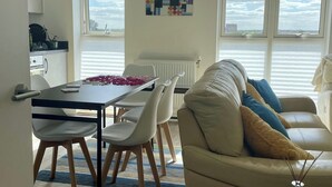 Ein modernes Wohnzimmer mit Sofa und Sesseln, einem Esstisch mit Rosenblättern und Meerblick durch große Fenster.