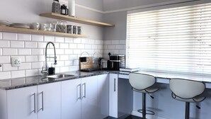 A modern kitchen with white cabinets, a granite countertop, a built-in oven, and a window with blinds. - 21 Cheviot Place (Cape Town)