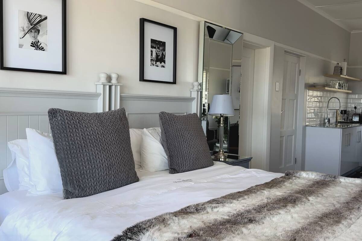 A hotel room with a bed, a mirror, a bedside table, and a view of the kitchen.