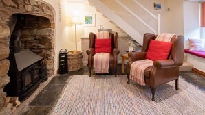 A cozy living room with a stone fireplace, two leather armchairs with red cushions, a woven basket, and a striped rug. - Gargoyle Cottage (St Agnes)